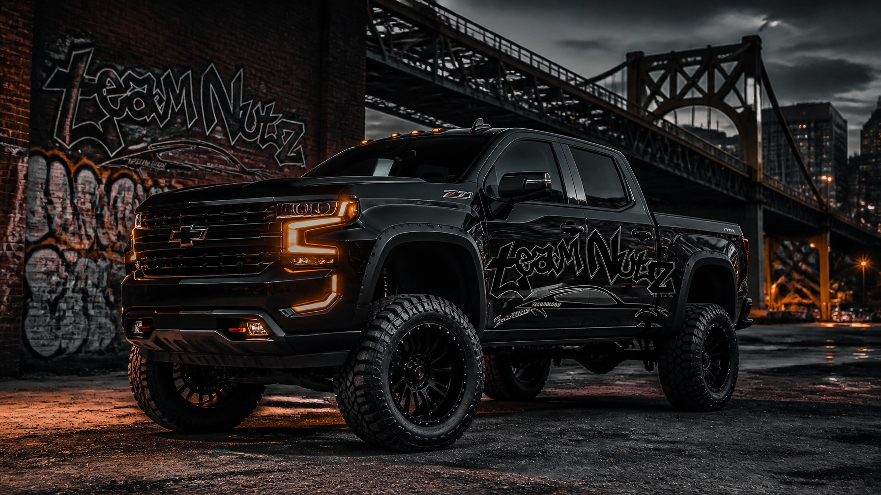 Black lifted Chevrolet pickup with orange accent lights and graffiti wrap under a urban bridge at dusk.
