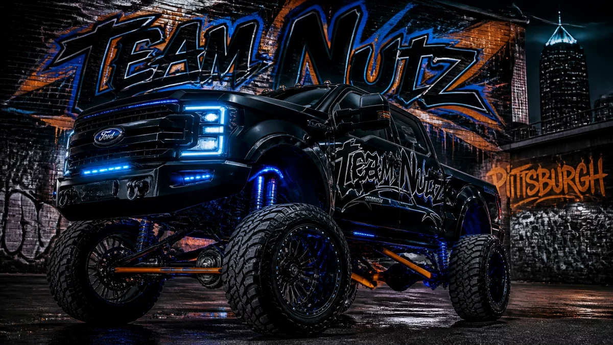 Monster black Ford monster truck with glowing blue lights and oversized tires in front of a graffiti wall that reads 'Team Nutz' and 'Pittsburgh' at night.