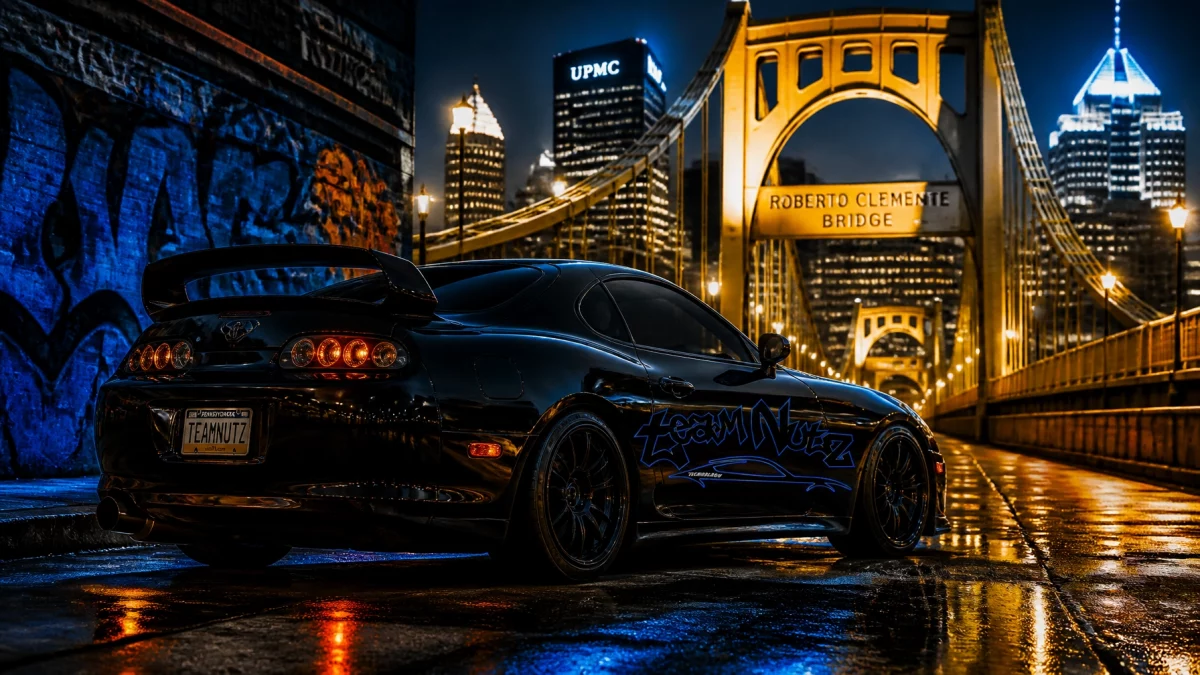 Night scene: a glossy black sports car with blue graffiti on the side sits on a wet street beneath the illuminated Roberto Clemente Bridge in Pittsburgh at night, reflections on the pavement.